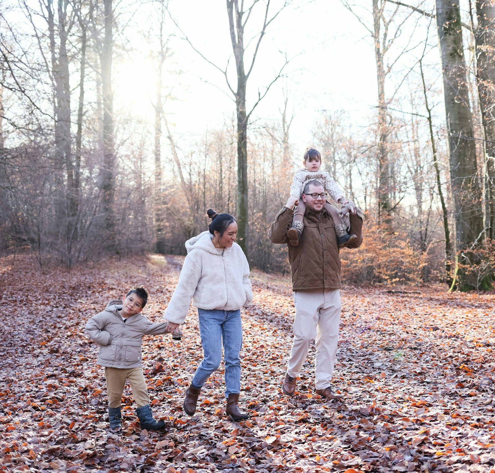 Familie mit zwei Kindern spaziert durch einen herbstlichen Wald, das kleinere Kind auf den Schultern des Vaters