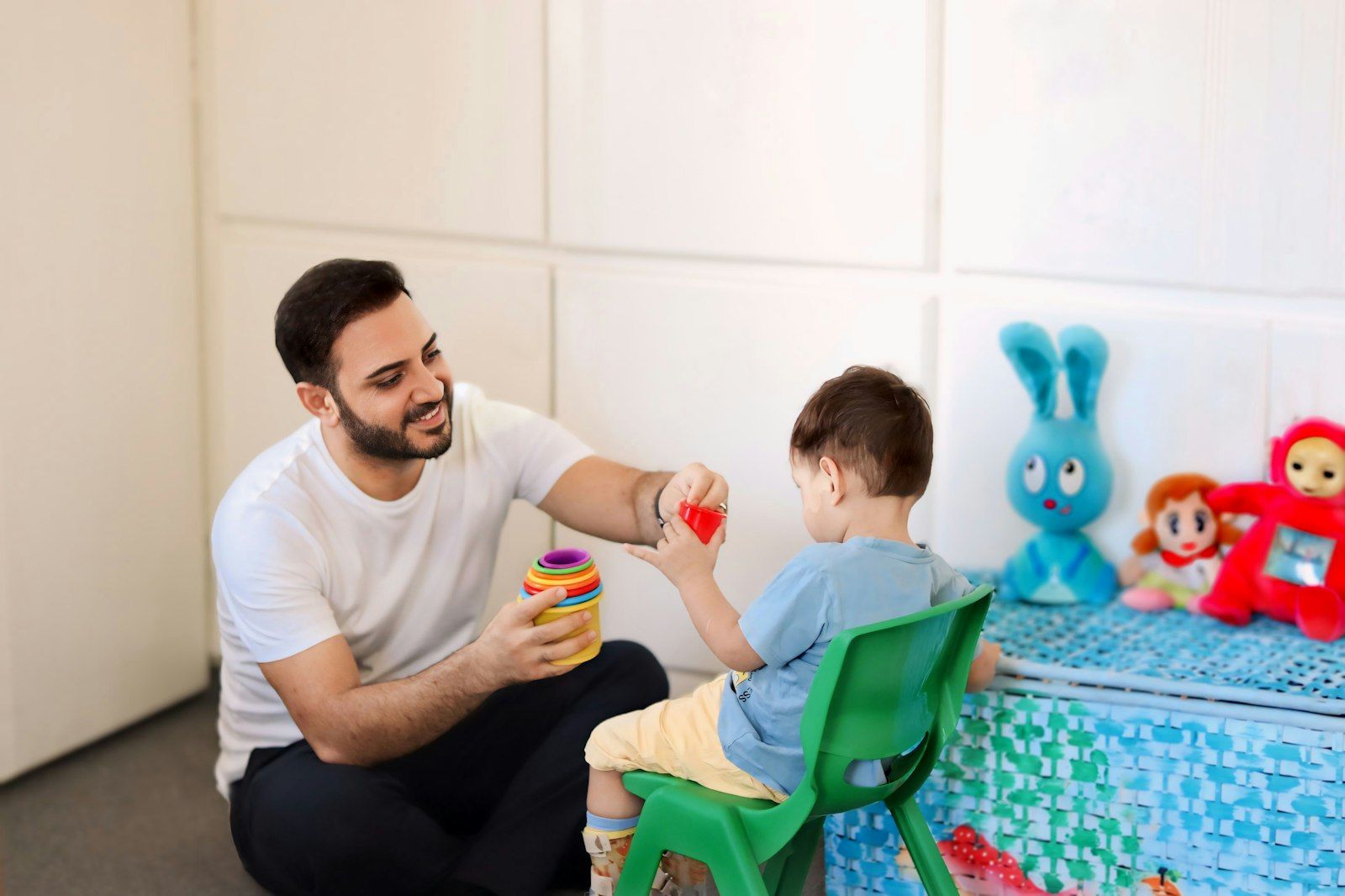 Mann spielt mit einem Kleinkind auf einem kleinen Stuhl mit bunten Stapelbechern